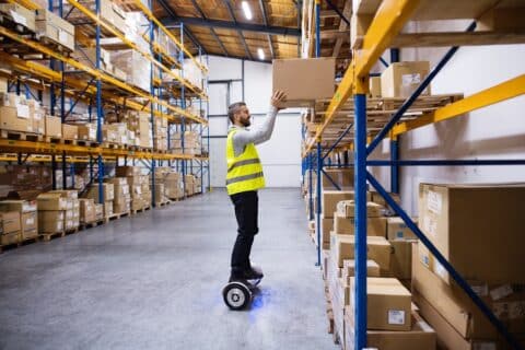 Un magasinier de sexe masculin sur un hoverboard, posant une grande boîte sur une étagère.