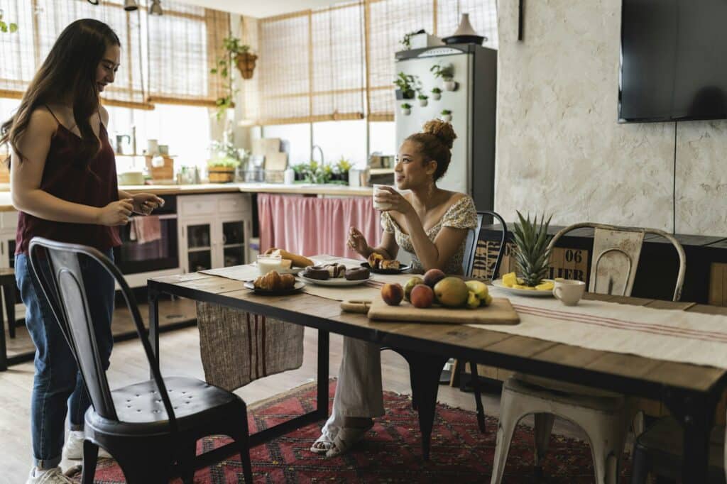 two multiracial women friends, having breakfast at table in kitchen, sharing flat, apartment
